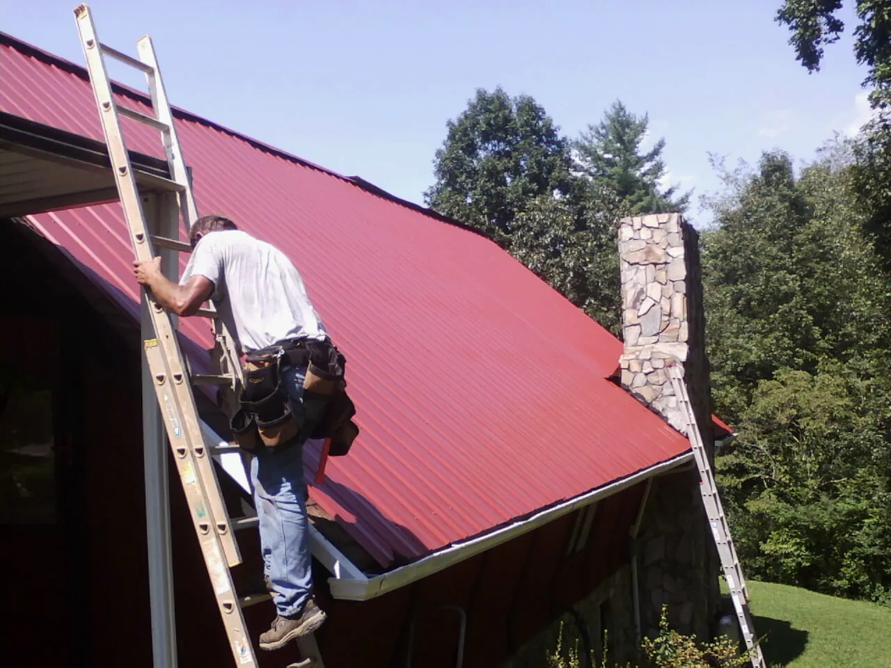 Roofer on a ladder beside a red metal roof and stone chimney — Soffit & Fascia Repair by Roofing Brothers in Deer Grove